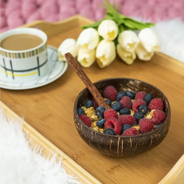 Kokosnuss-Schale gefüllt mit Müsli und Beeren steht auf einem Tablett, auf dem sich eine Tasse Kaffee und ein Strauß Tulpen befinden